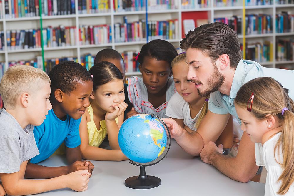 Eine multikulturelle Schulklasse ist in einer Bücherei um einen kleinen Globus versammelt.
